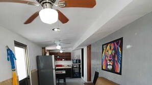 Ein Wohnzimmer mit einer Couch, einem Deckenventilator, einer Küche im Hintergrund und einem farbenfrohen Bild an der Wand.