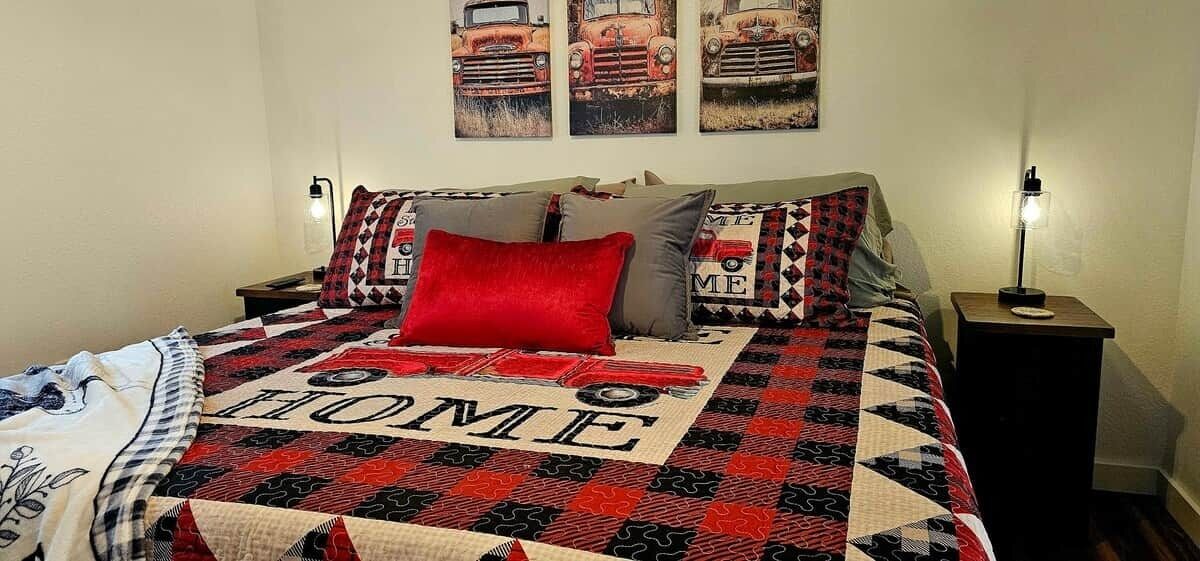 Uma cama com colcha decorada com um carro e a palavra 'HOME', duas luminárias de cabeceira e uma parede com três quadros de tema automotivo.