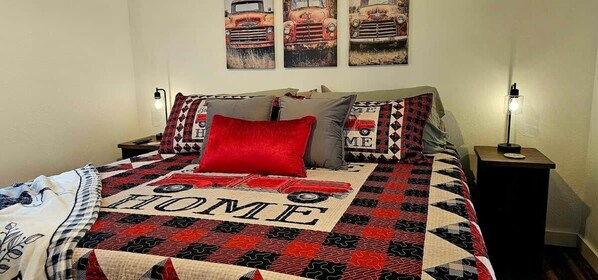 A bed with a quilt featuring a car and the word 'HOME,' two bedside lamps, and a wall with three car-themed paintings.