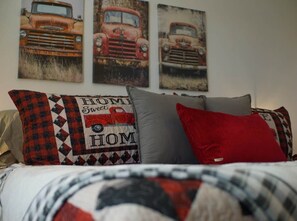 Una cama con una manta y almohadas estampadas, una pared con tres carteles de autos antiguos, y una vista parcial de una ventana.