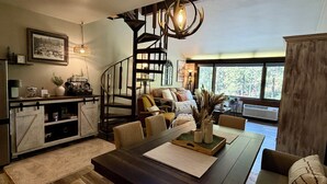 Uma sala espaçosa com uma mesa de jantar de madeira, um conjunto de sofás, uma escada em espiral e uma grande janela com vista para o exterior.