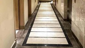 Um corredor comprido e estreito com chão de azulejos, portas de madeira e uma parede de mármore em uma lateral.