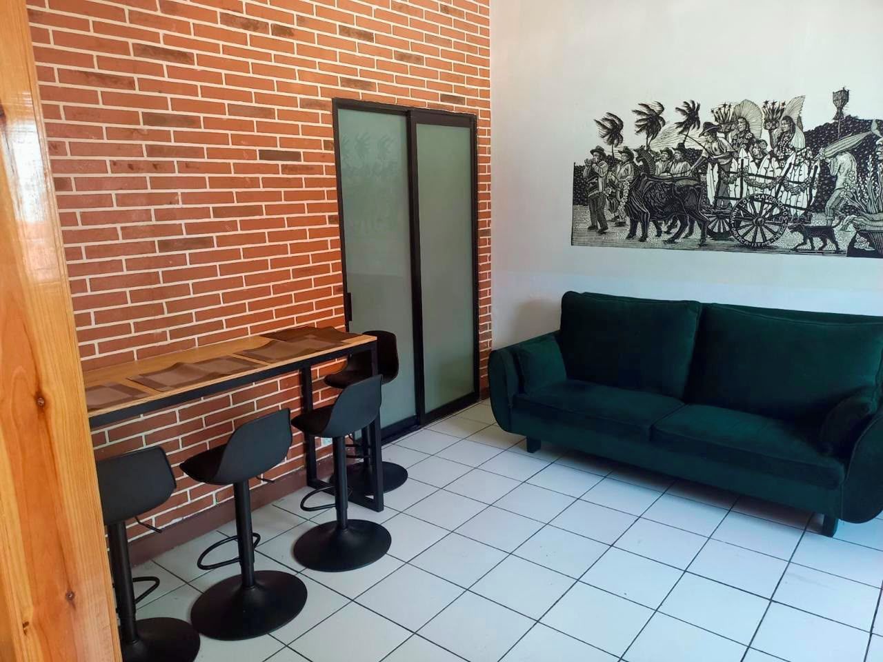 Una habitación con pared de ladrillo, sofá verde, mesa de madera con sillas y una obra de arte enmarcada en la pared.