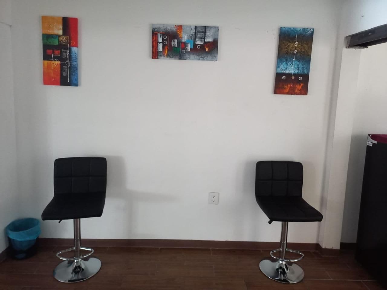 Two black chairs with metal bases, a blue trash bin, and three abstract paintings on the wall.