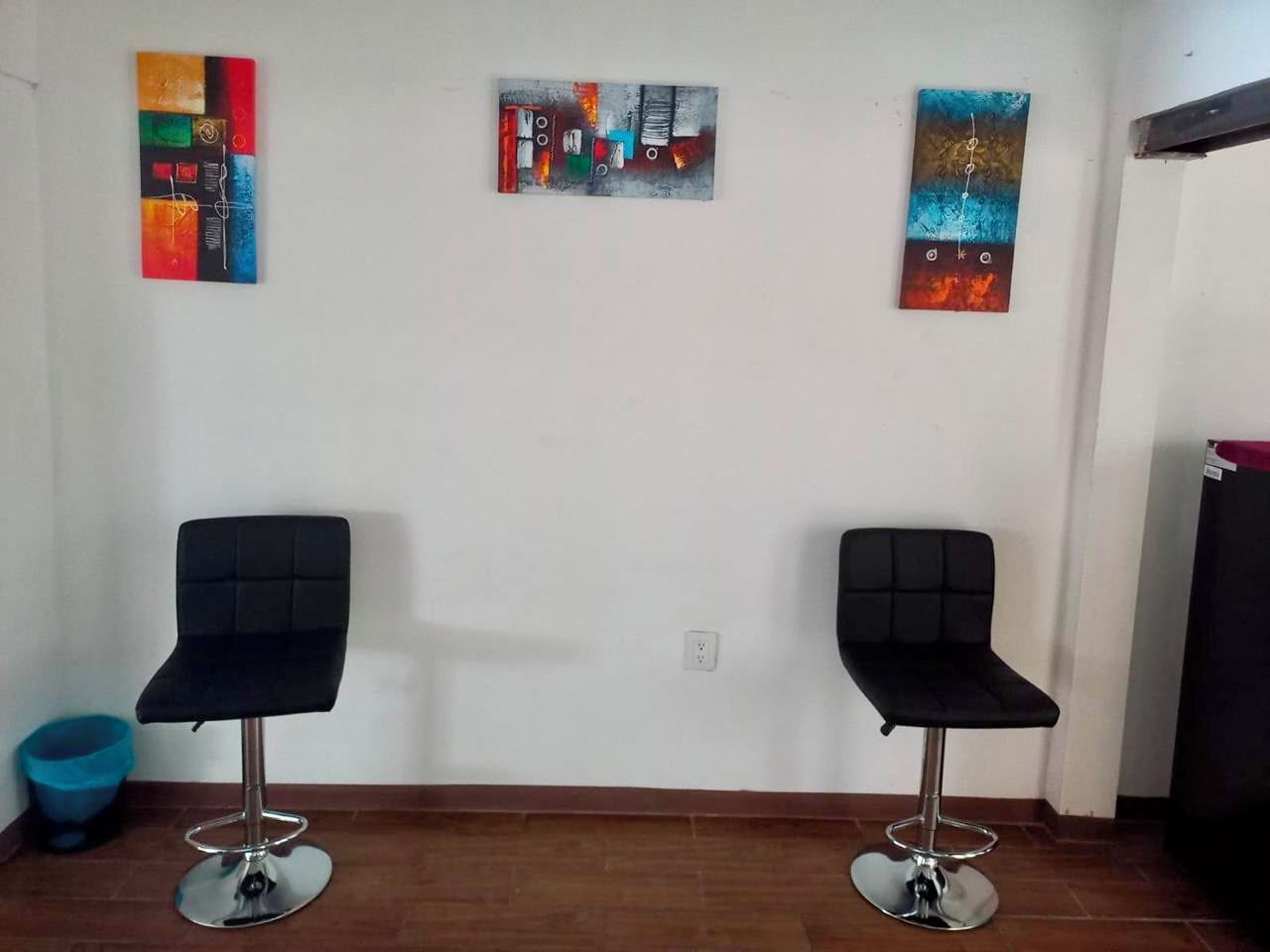 Una habitación con dos sillas negras, un cesto de basura azul y tres cuadros abstractos en la pared.
