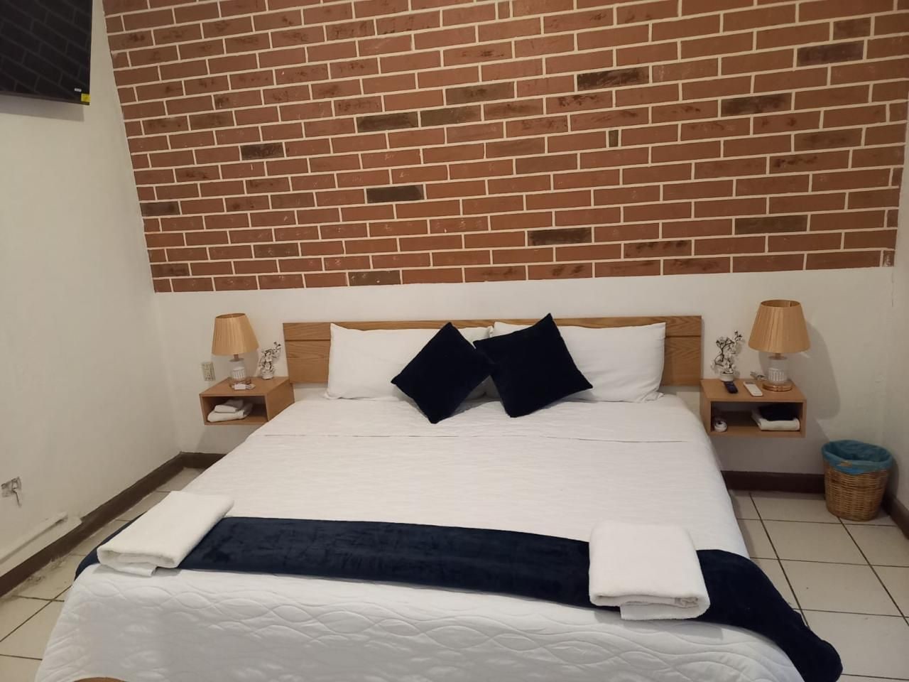 A hotel room with a brick-patterned headboard, a neatly made bed with white and dark blue linens, two bedside lamps, and a small wicker basket.