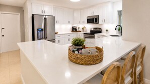 Uma cozinha moderna com armários brancos, eletrodomésticos de aço inoxidável, uma ilha central com cesta de vime e cadeiras de madeira.