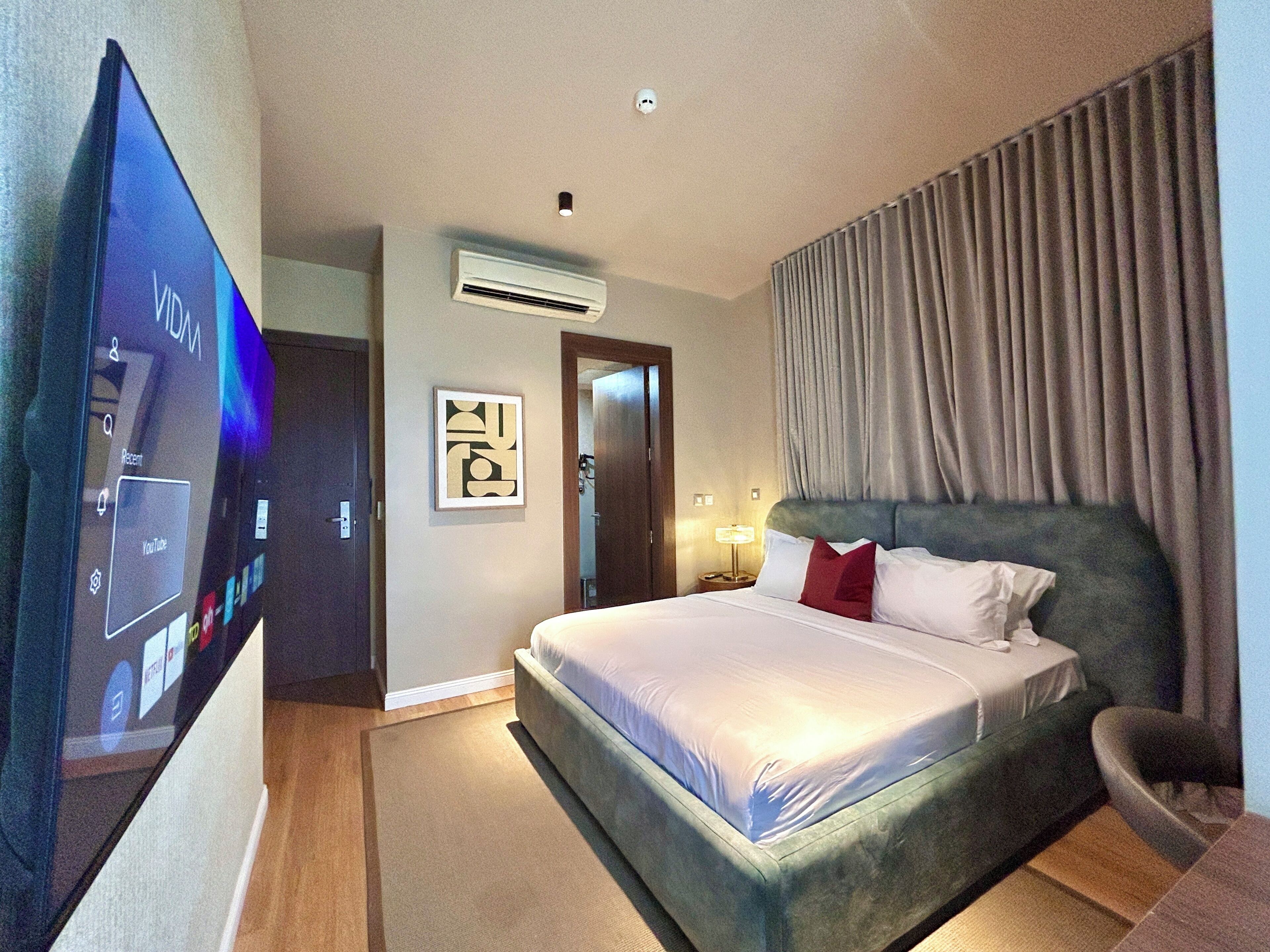 Quarto de hotel com uma televisão de tela plana grande, uma cama com cabeceira, ar-condicionado, um quadro na parede e uma janela com cortinas.