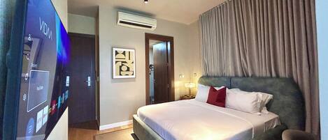 A hotel room with a large flat-screen TV, a bed with a headboard, an air conditioning unit, a framed picture on the wall, and a window with curtains.