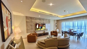 A modern living room with a flat-screen TV, comfortable seating, a dining area, and a large window with curtains.