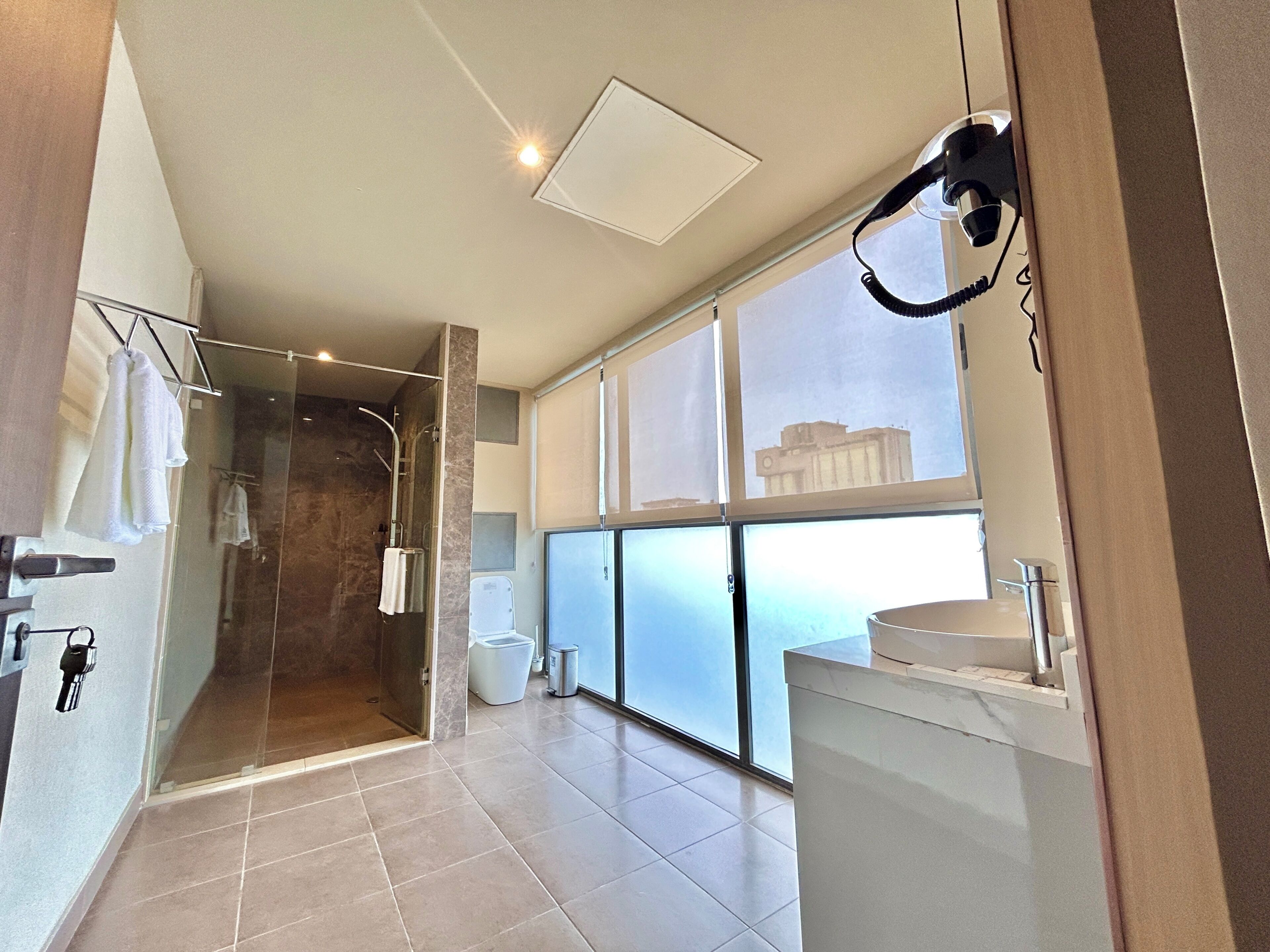 A modern bathroom with a glass shower enclosure, a large window with a city view, and a hairdryer mounted on the wall.