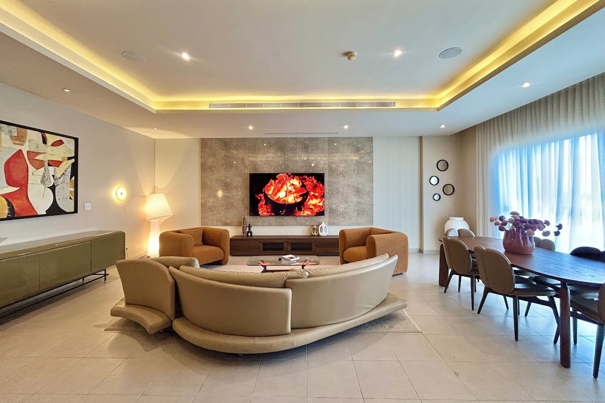 A modern living room with a curved sofa, a flat-screen TV, a dining table with chairs, and a large window with curtains.