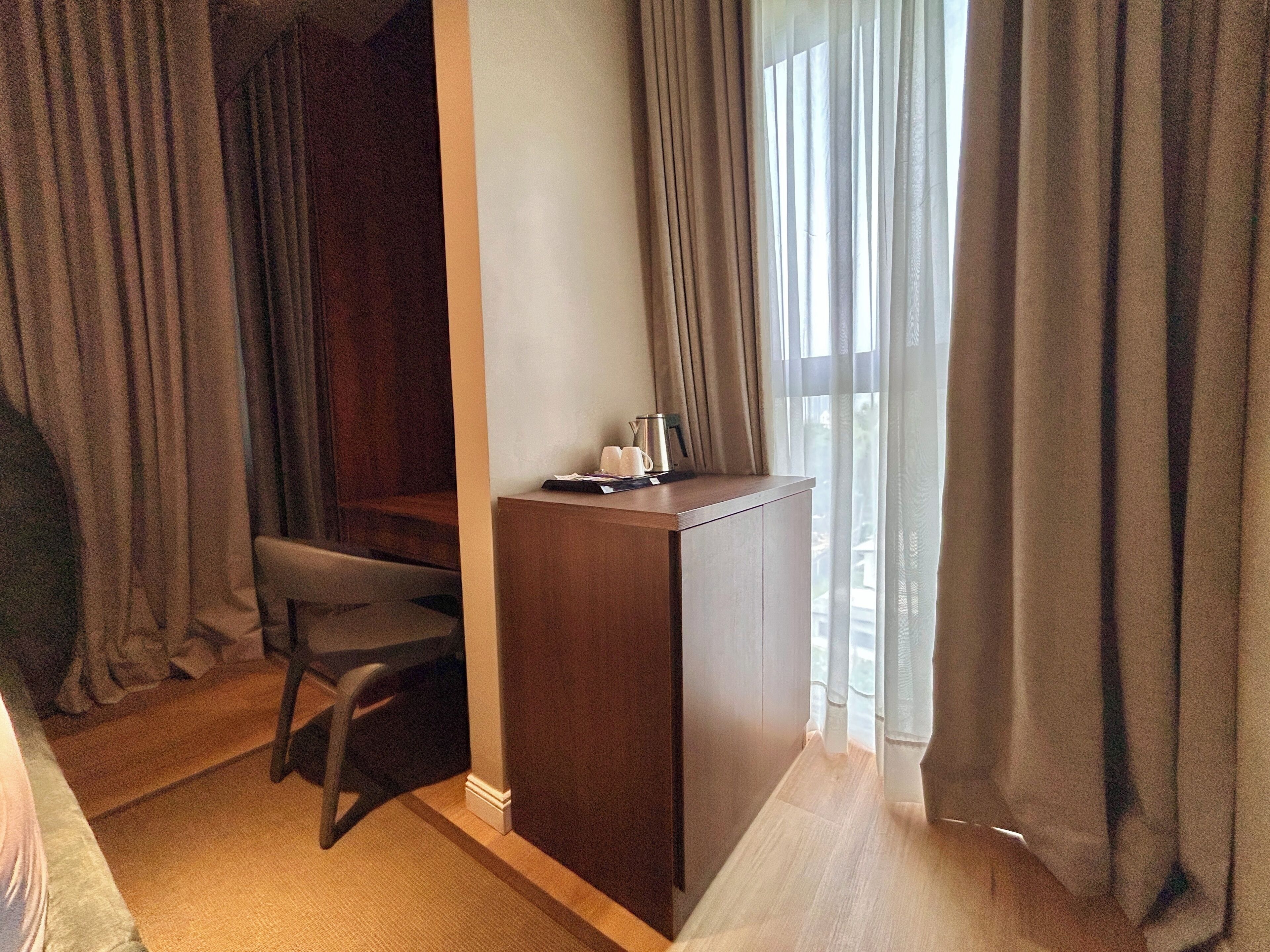 A hotel room with a wooden desk, a chair, a window with curtains, and a view of the outside.