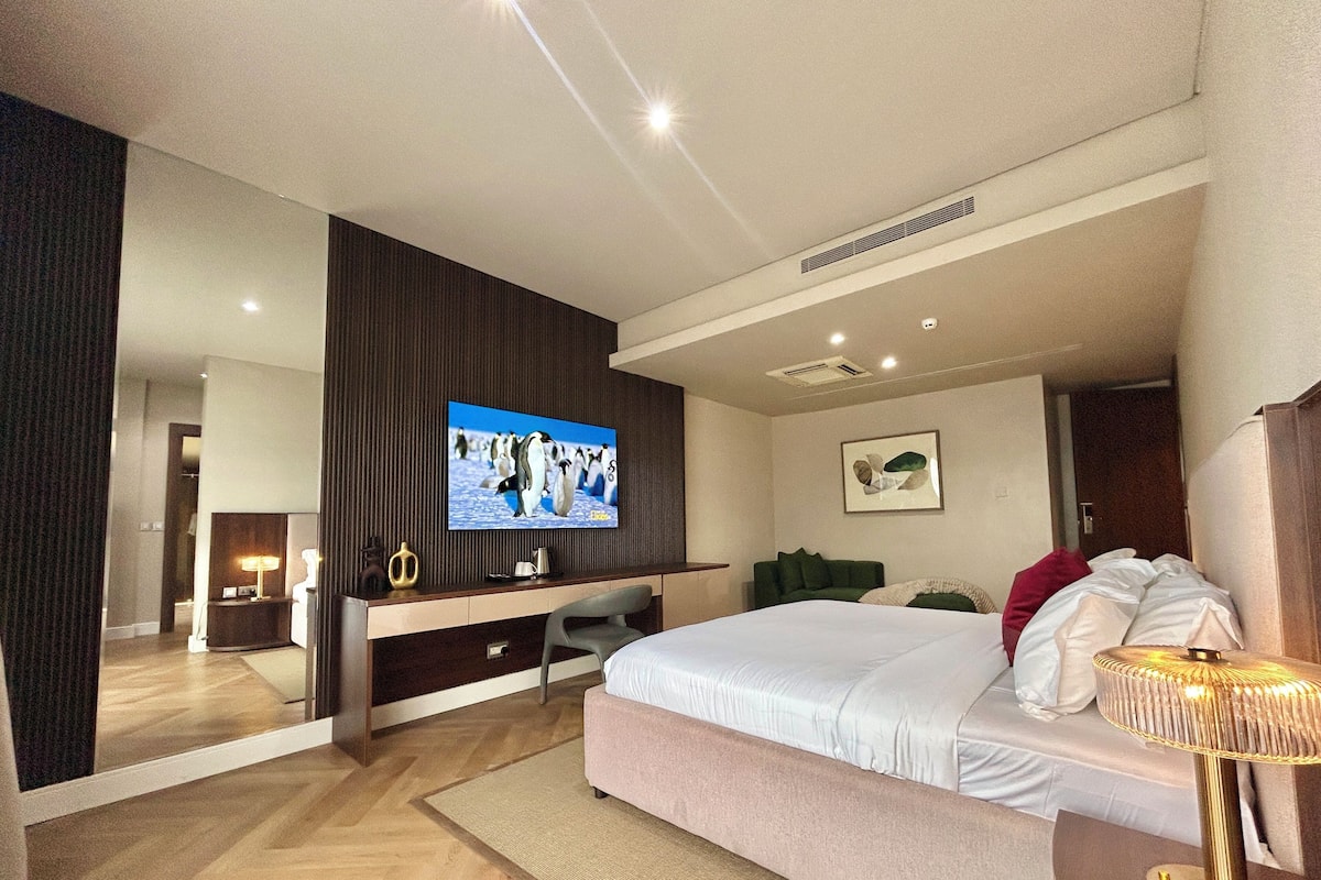 A modern hotel room with a large bed, a desk with a computer, a television, and a mirror.