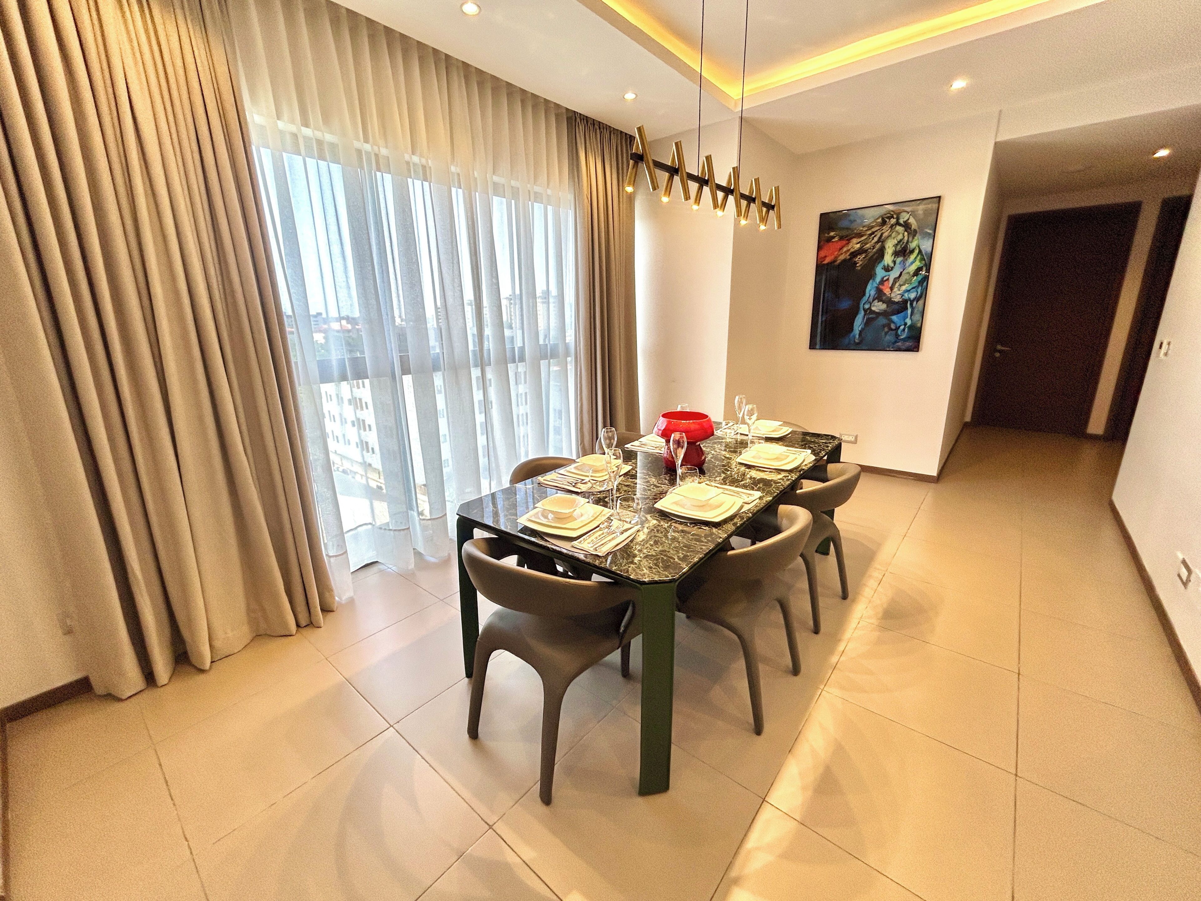 A dining area with a table set for four, a modern chandelier, and a city view through large windows.