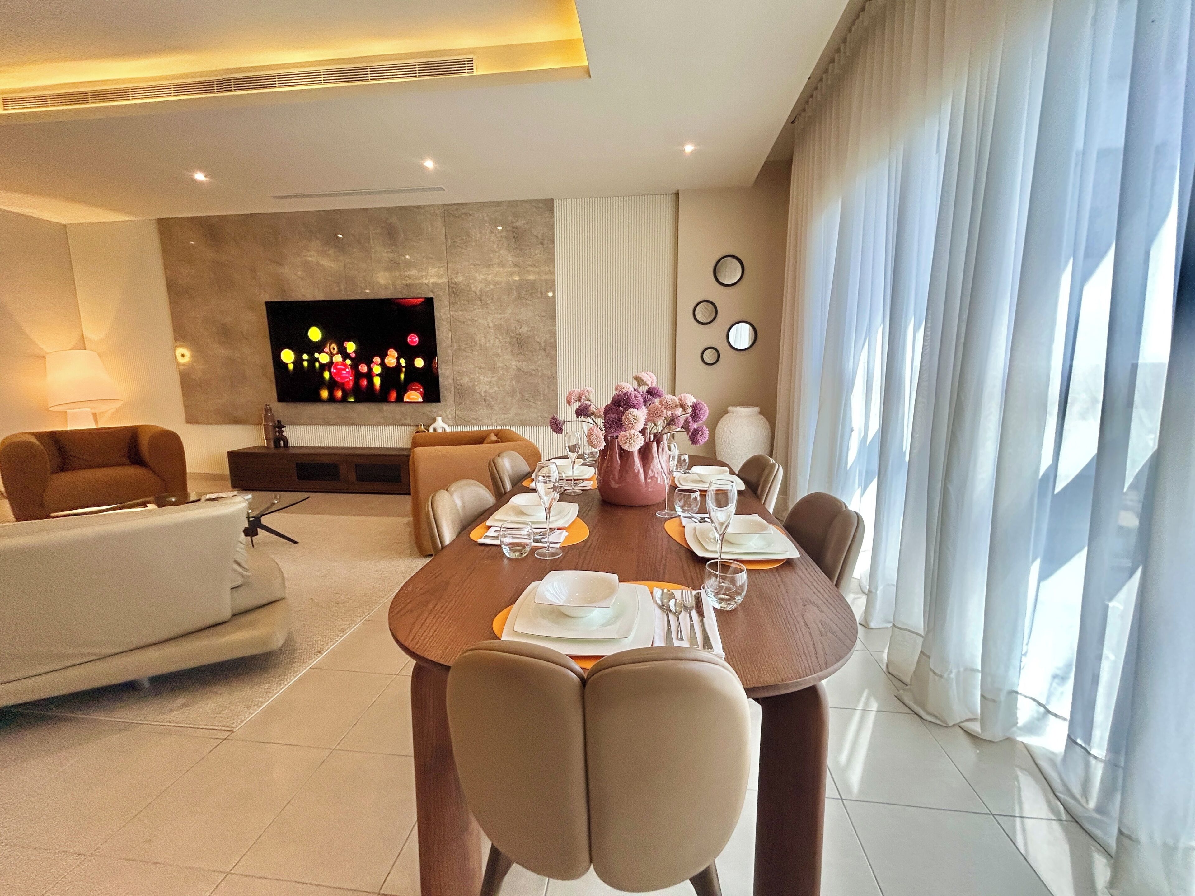 A modern living room with a dining area, a sofa, a TV, and a large window with curtains.