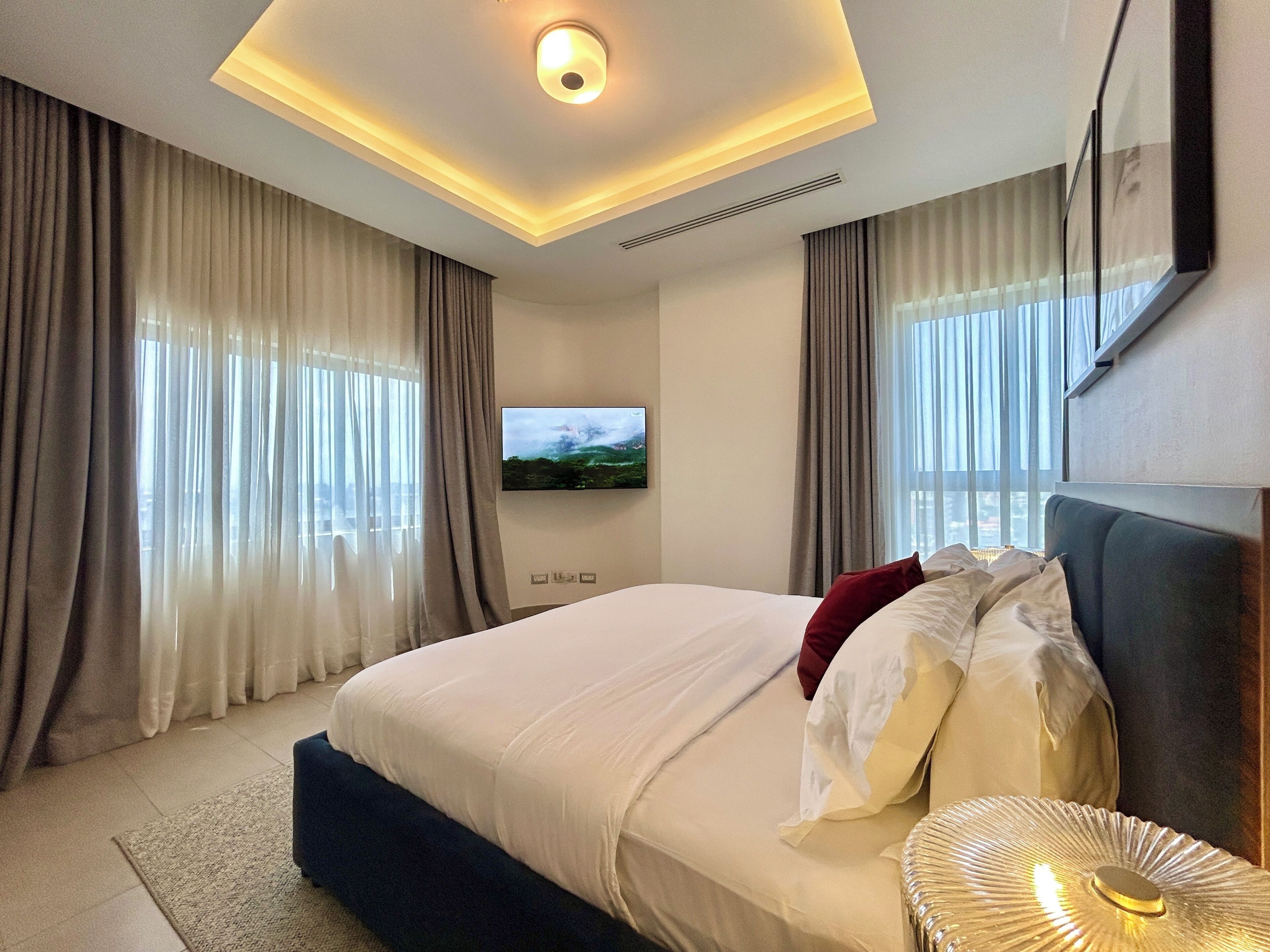 A modern hotel room with a large bed, a flat-screen TV, and a view of the city through the windows.