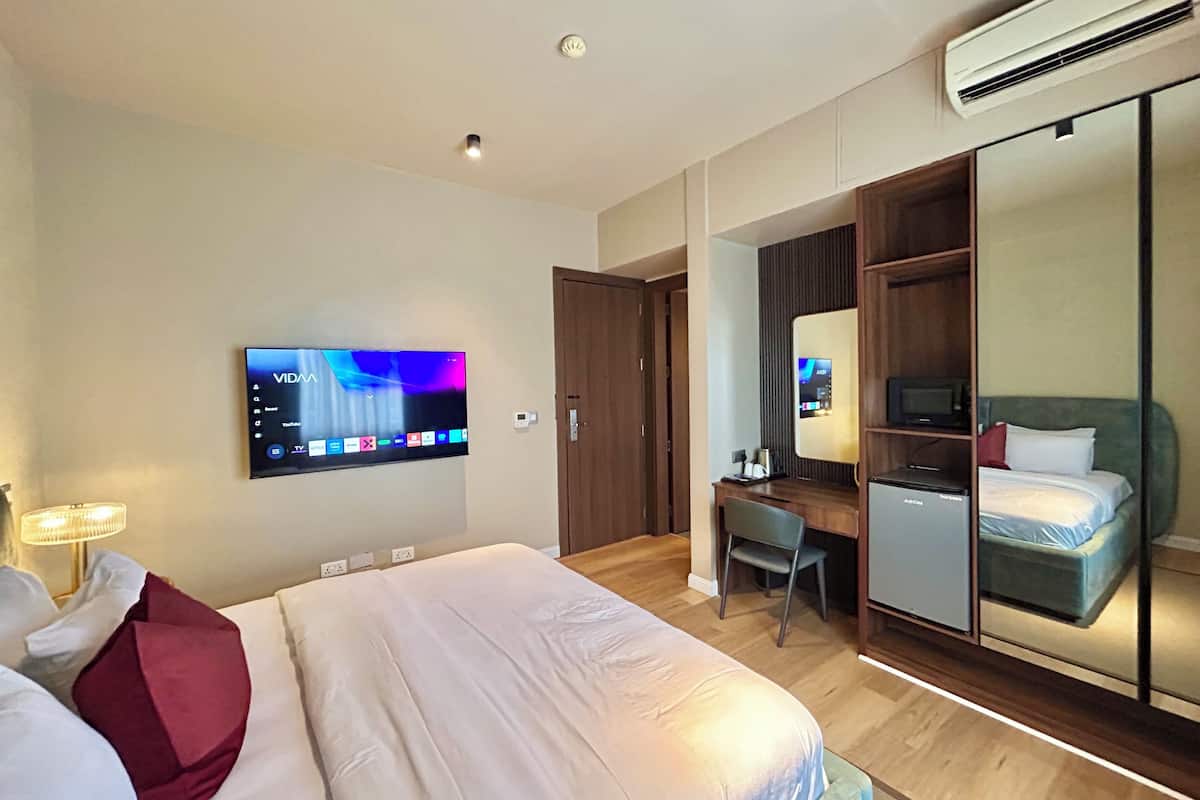 A hotel room with a bed, a desk, a chair, a TV mounted on the wall, a wardrobe, and a mirror.