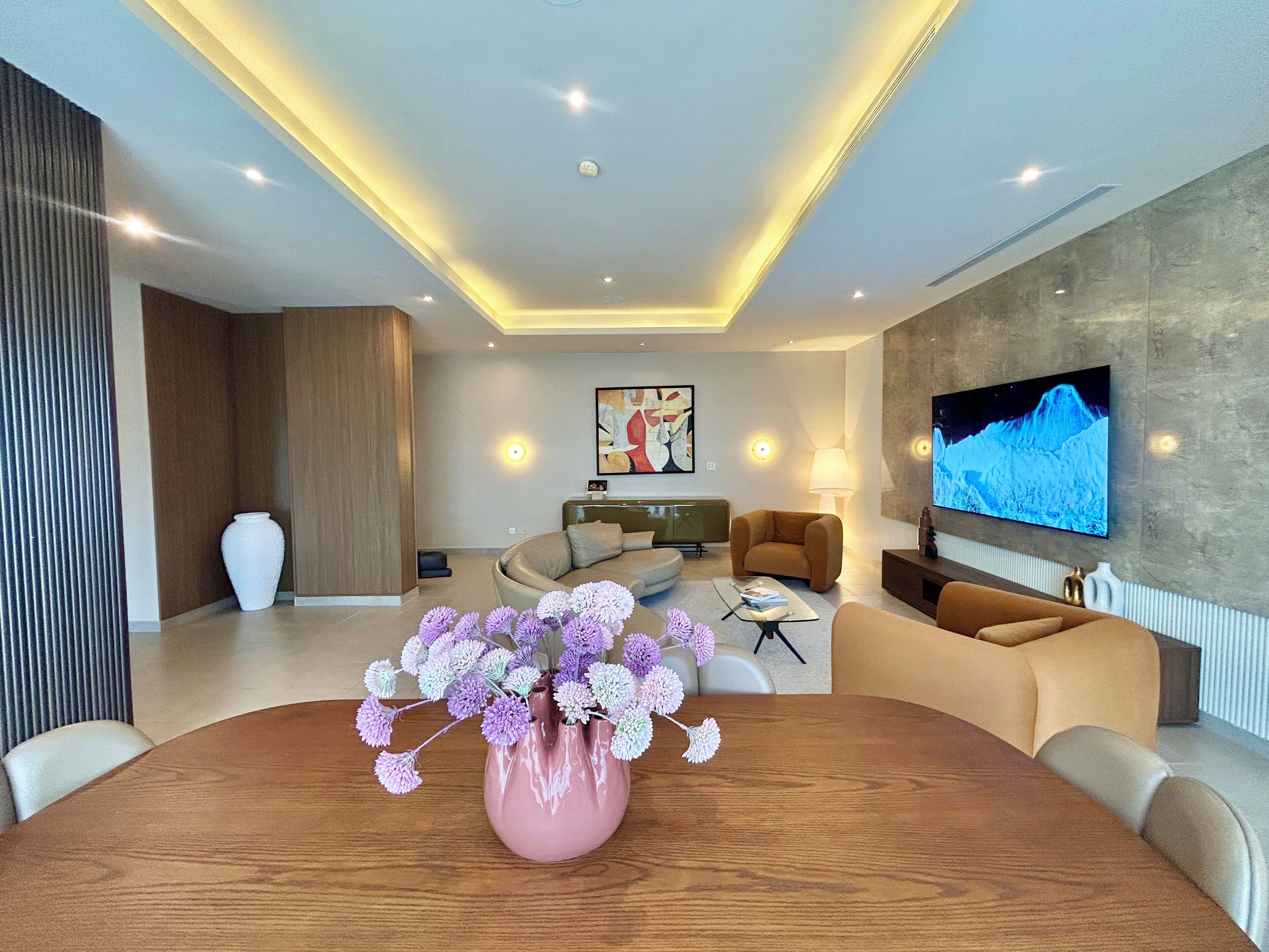 Ein modernes Wohnzimmer mit einem Holzessttisch, einer rosa Vase mit Blumen, einer Couch, einem Fernseher und einem Wandbild.