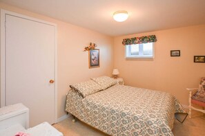 A bedroom with a bed, a nightstand, a chair, a door, and a window with a floral curtain.