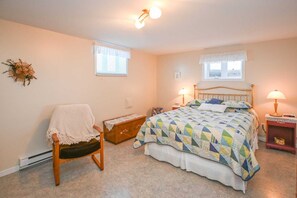 A bedroom with a bed, two bedside lamps, a chair with a blanket, a small wooden chest, and a window with a view.