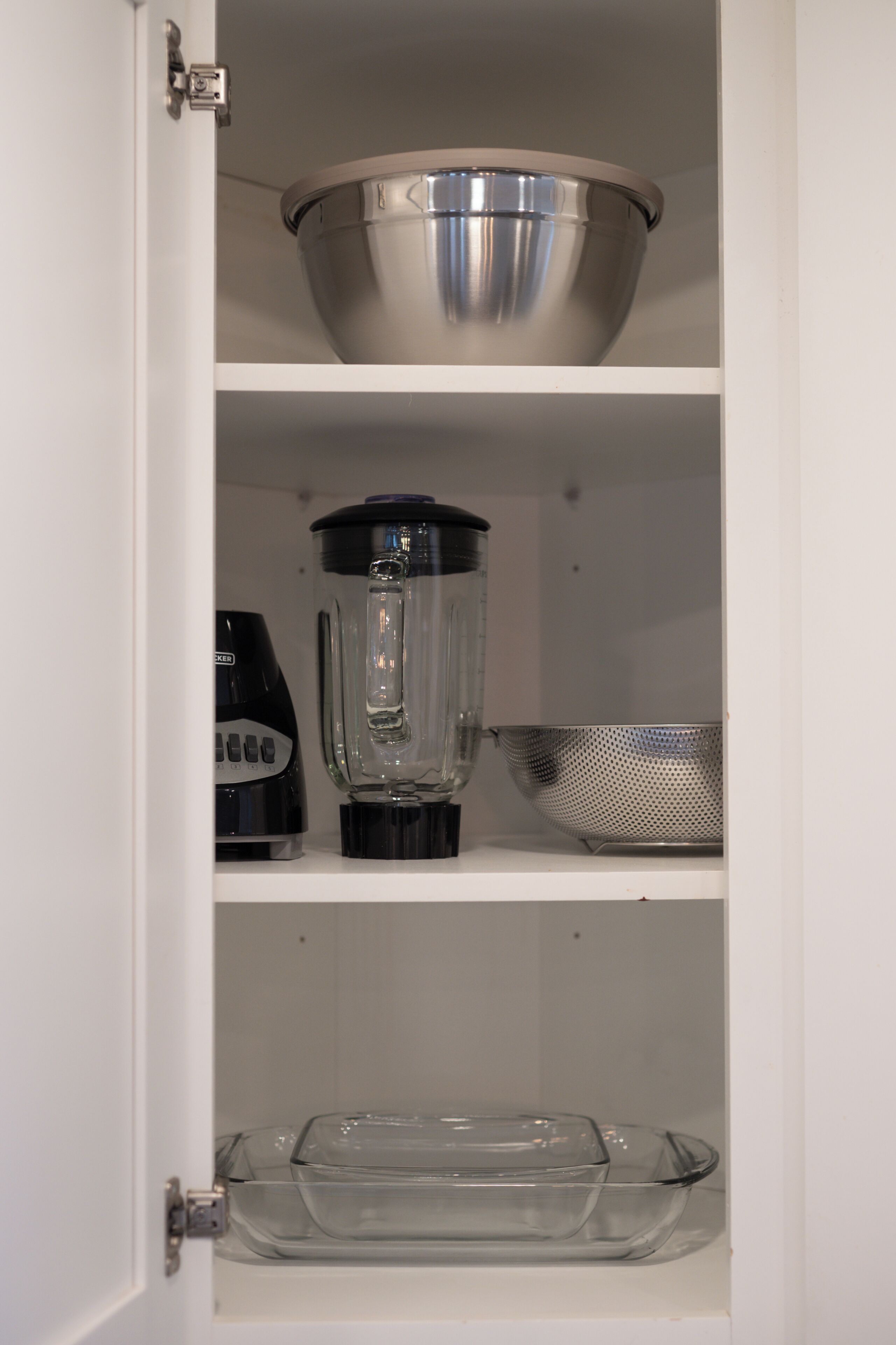 Un meuble de cuisine avec un saladier en acier inoxydable sur l’étagère supérieure, un mixeur sur l’étagère du milieu et un plat de cuisson en verre sur l’étagère inférieure.