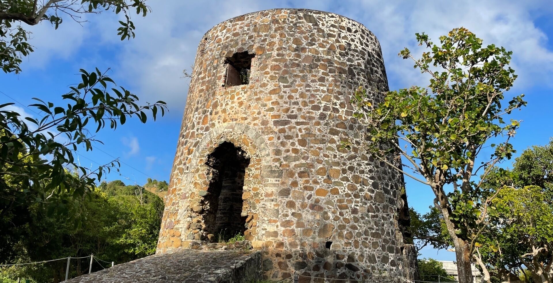 A stone tower with a central arched opening, surrounded by trees and greenery.