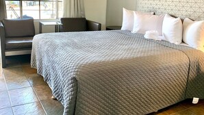 A hotel room with a large bed, a small table, a chair, and a window with curtains.