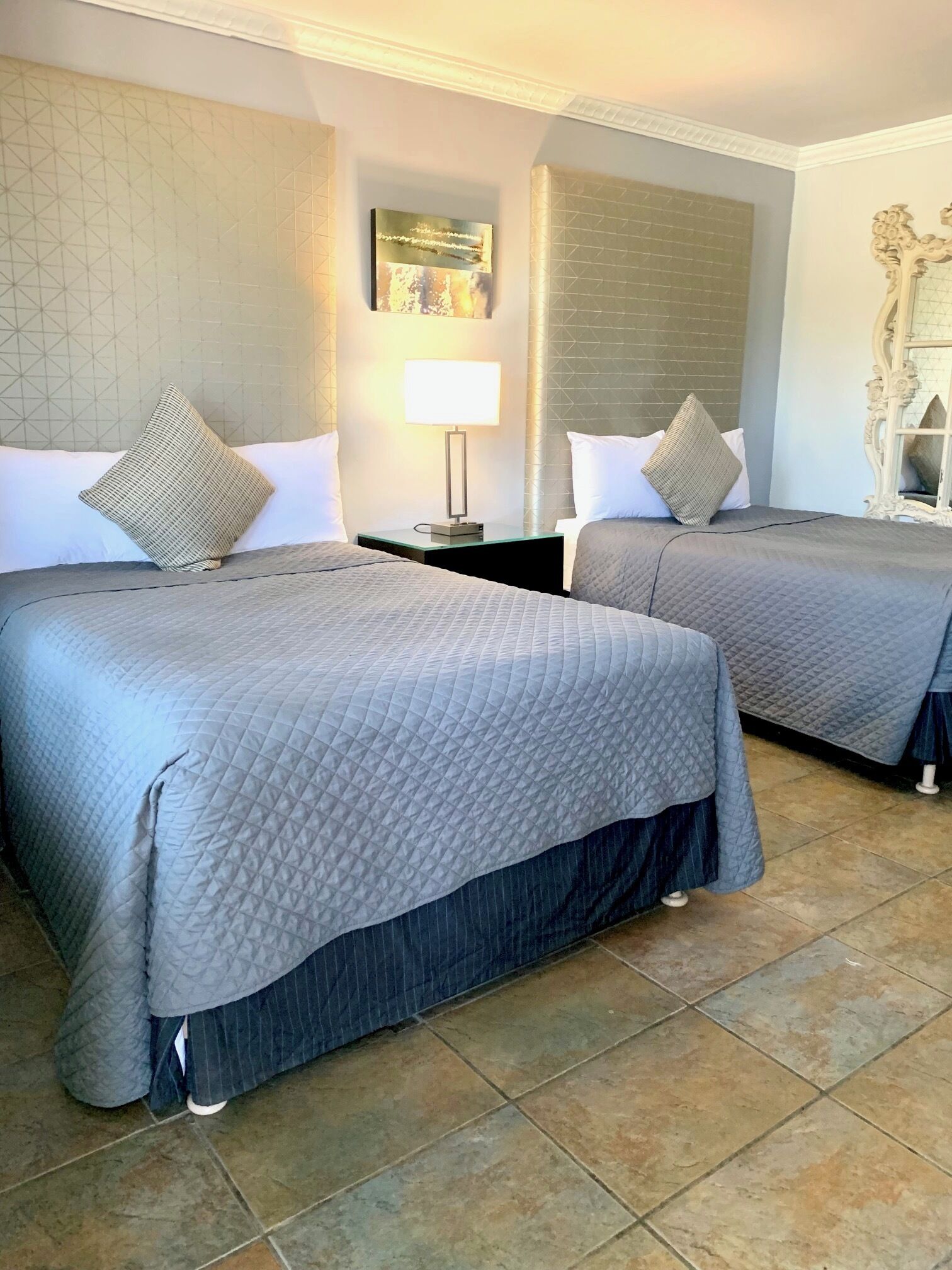 Habitación de hotel con dos camas, una mesita de noche con lámpara, un espejo decorativo y una pared con estampado geométrico.