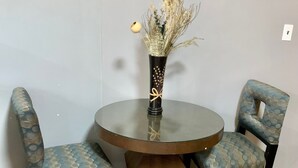 Una mesa redonda de cristal con dos sillas tapizadas, un jarrón con flores secas y un interruptor de luz en la pared.