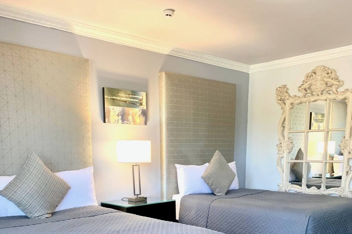 Ein Hotelzimmer mit zwei Betten, einem Nachttisch mit Lampe, einem Zier-Spiegel und einem Gemälde an der Wand.