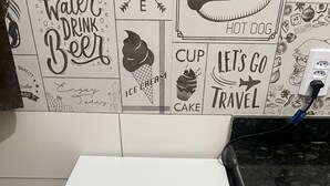 A compact white Midea refrigerator with a 10-year warranty sticker, a power outlet on the countertop, and a tiled backsplash with various food-related illustrations.