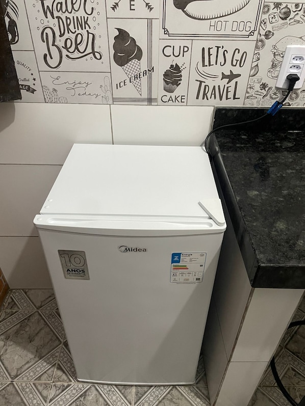 A compact white Midea refrigerator with a 10-year warranty sticker, a power outlet on the countertop, and a tiled backsplash with various food-related illustrations.