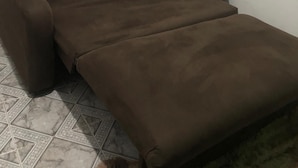 A small, single-bed room with a brown bedspread, a single pillow, and a window with white curtains.