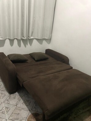 Una habitación pequeña con cama individual, colcha marrón, una almohada y ventana con cortinas blancas.