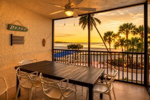 Ein Balkon mit einem Holztisch und weißen Stühlen, einem Deckenventilator und Blick auf den Strand bei Sonnenuntergang.