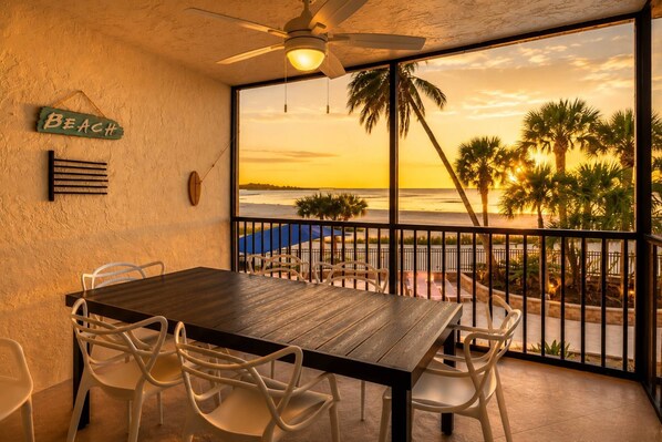 Un balcon avec une table en bois et des chaises blanches, un ventilateur de plafond et une vue sur la plage au coucher du soleil.