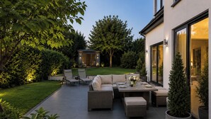Une terrasse extérieure aménagée avec un ensemble de canapés, une table à manger et des chaises, le tout entouré de verdure et d’une petite construction.