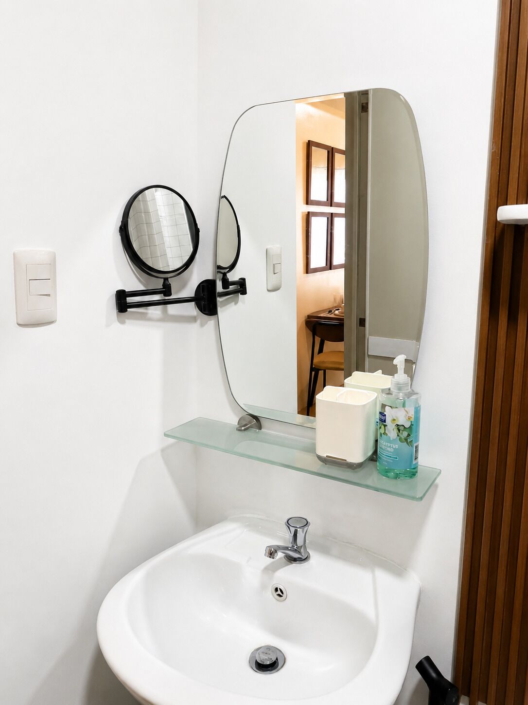 A bathroom with a white sink, a round mirror, a glass shelf with toiletries, and a wooden door.
