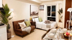 A modern living room with a brown sofa, a dining table set for four, and a large window with curtains. - Cozy 29F Avida Riala IT Park Sea & City View (Cebu City)