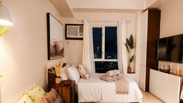 A modern bedroom with a bed, a sofa, a TV, and a window with a city view at night. - Cozy 29F Avida Riala IT Park Sea & City View (Cebu City)