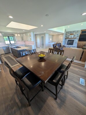 Amplia sala de estar con una gran mesa de madera para comer, asientos cómodos, una chimenea de piedra y una cocina moderna al fondo.