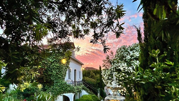 Un jardin avec une fontaine, un bâtiment blanc orné d’arcades et un coucher de soleil en arrière-plan.