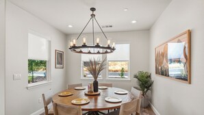 A dining room with a round wooden table, six chairs, a chandelier, and two windows with views. - Amazing new home- All the Amenities! (Murphys)