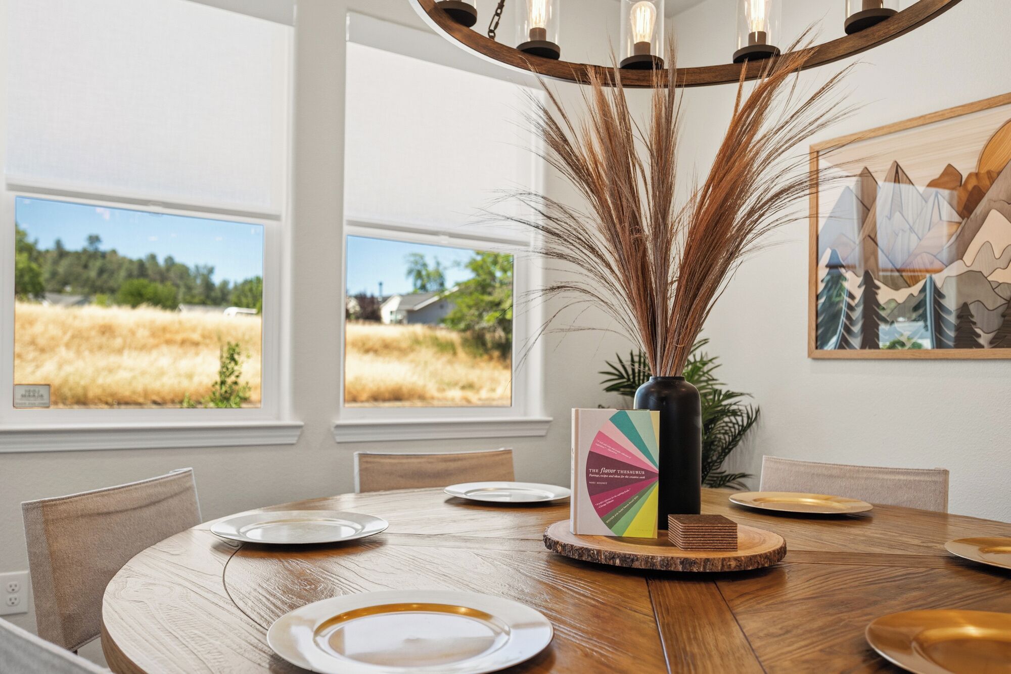 A dining area with a round wooden table, chairs, a vase with tall grasses, a framed picture, and a menu stand.