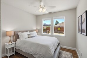 A bedroom with a bed, nightstand, lamp, ceiling fan, and two windows with a view of the backyard.