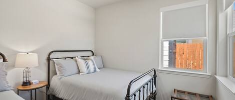 A bedroom with a bed, bedside table, lamp, window, and a small wooden stool.