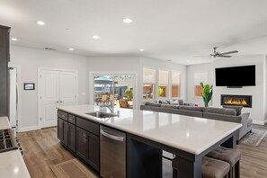 A modern kitchen with a central island, a dining area with a sofa, a TV mounted on the wall, and a fireplace.