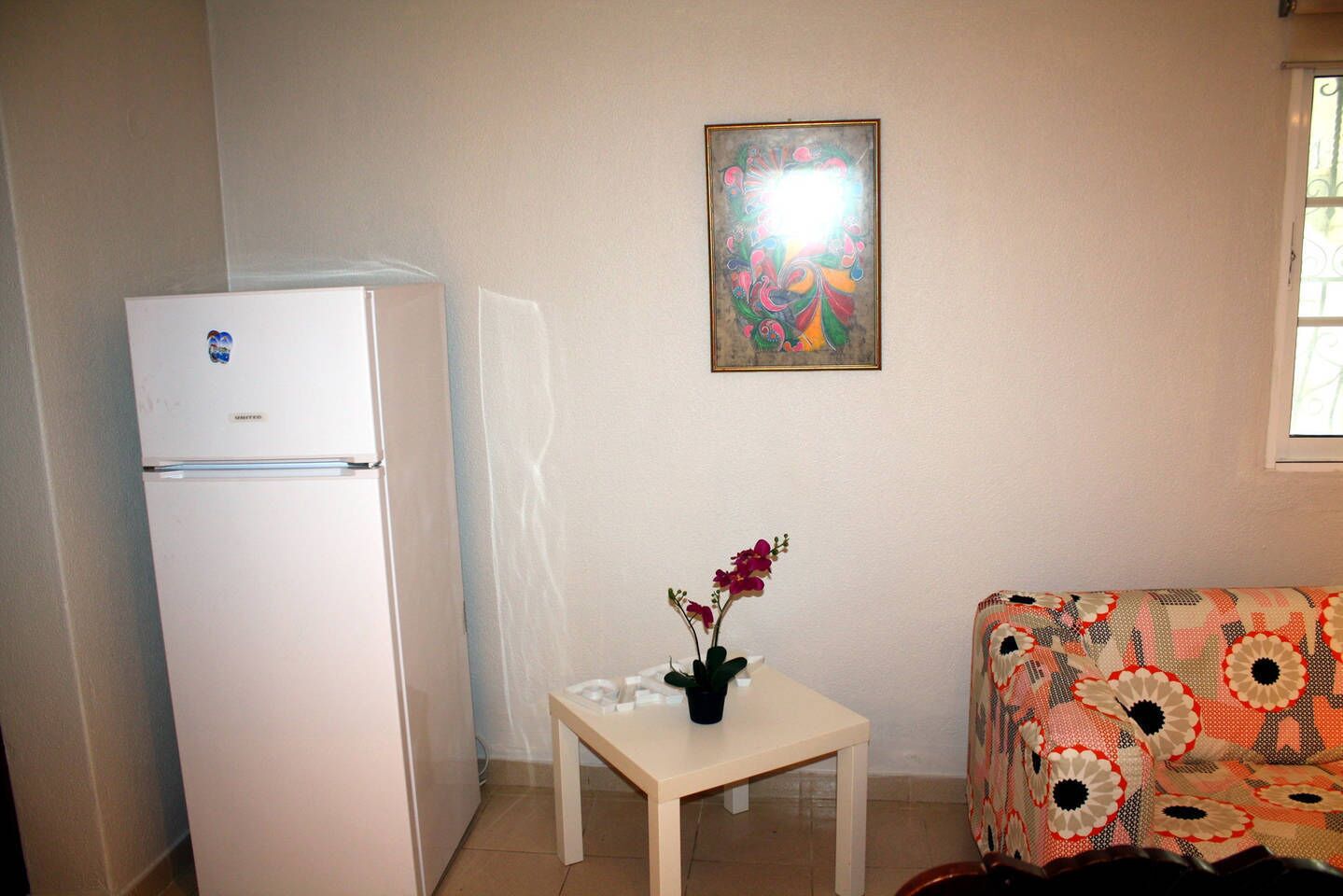 A small room with a white refrigerator, a table with a potted plant, a colorful patterned sofa, and a wall with a framed artwork.