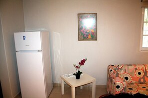 Ein kleines Zimmer mit einem weißen Kühlschrank, einem Tisch mit einer Topfpflanze, einem farbenfrohen gemusterten Sofa und einer Wand mit einem gerahmten Kunstwerk.
